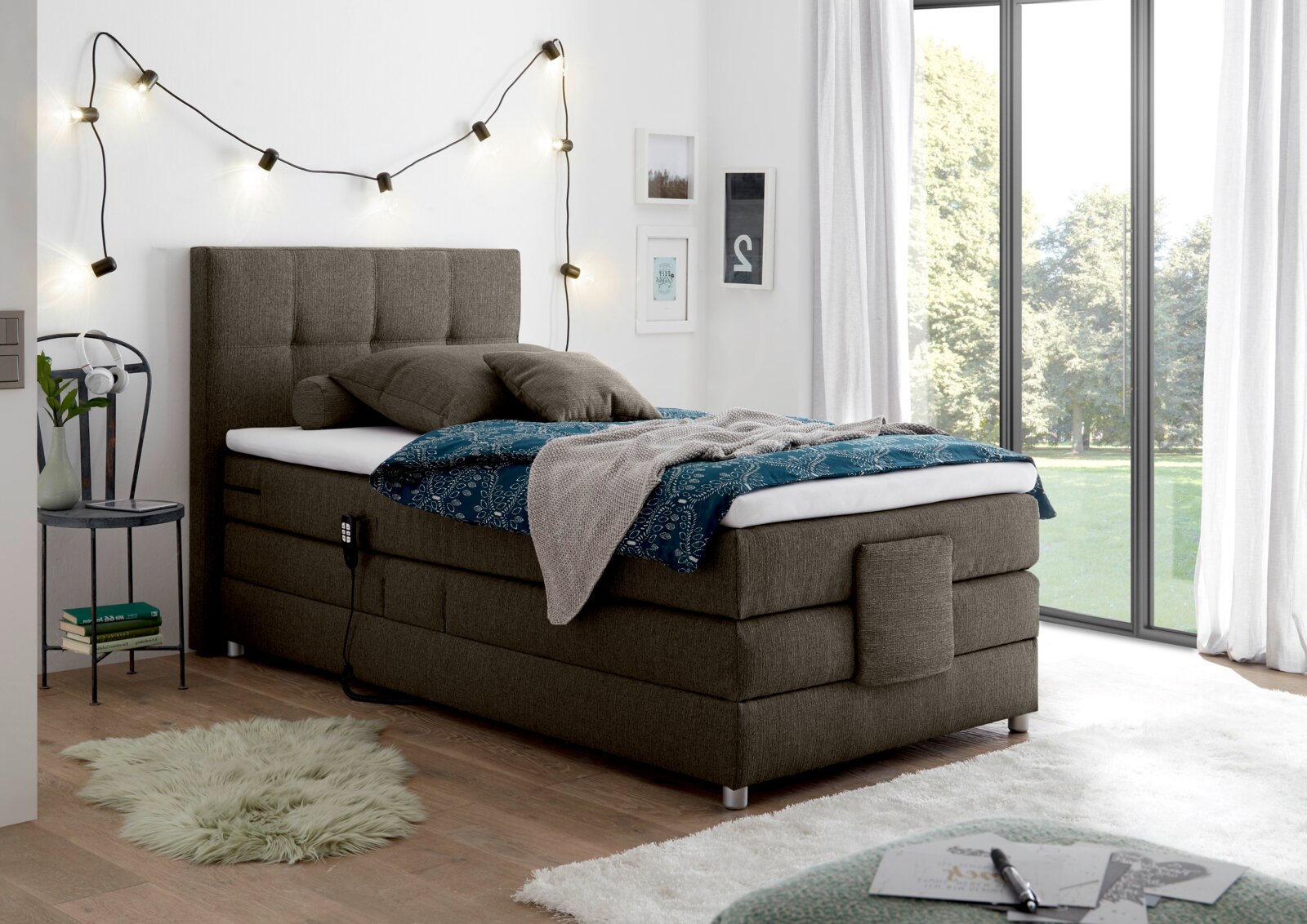 Esposa Boxspringbett MANOLO Boxspringbett in modernem Schlafzimmer, seitliche Perspektive, mit grauem Stoffbezug, dekoriert mit Kissen und Decke, neben einem großen Fenster.