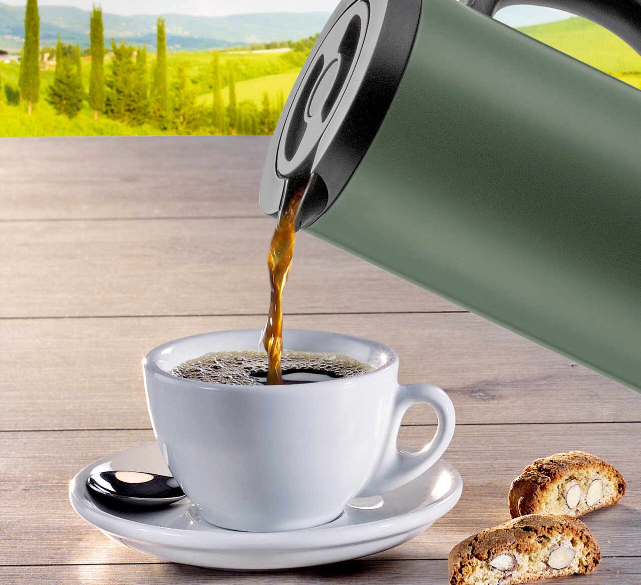 Grüne Isolierkanne beim Einschenken von Kaffee in eine weiße Tasse, mit Keksen auf einem Holztisch und einer Landschaft im Hintergrund, seitliche Perspektive.