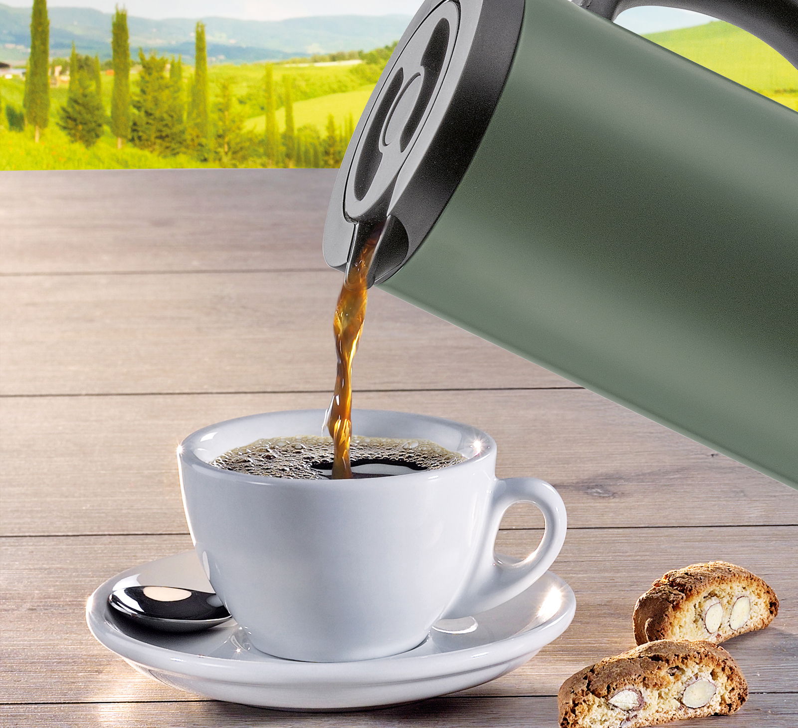 Cilio Isolierkanne FIRENZE 1L Grüne Isolierkanne beim Einschenken von Kaffee in eine weiße Tasse, mit Keksen auf einem Holztisch und einer Landschaft im Hintergrund, seitliche Perspektive.