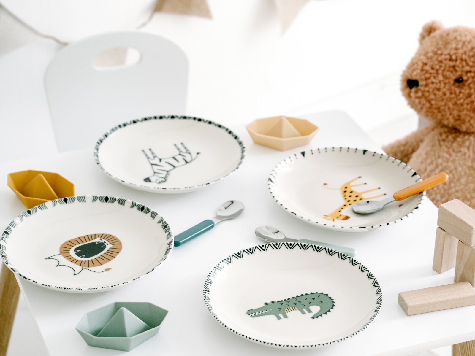 creaTable Teller-Set 4-tlg SAFARI Schräg von oben fotografiertes Kinder-Geschirrset auf weißem Tisch: vier Teller mit Tiermotiven (Löwe, Zebra, Giraffe, Krokodil), dazu kleine Schälchen und Kinderlöffel, daneben ein Teddybär.