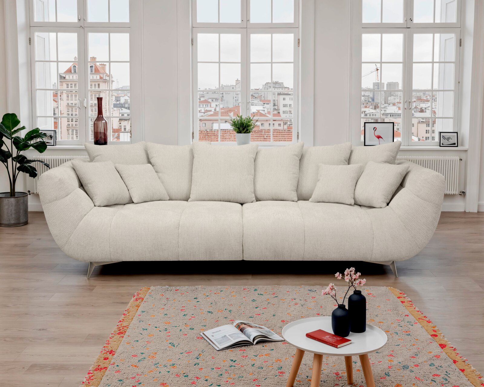Helles, cremefarbenes Designer-Sofa mit vielen Kissen und schmalen Metallfüßen im modernen Wohnzimmer vor großen Fenstern, Frontalansicht.