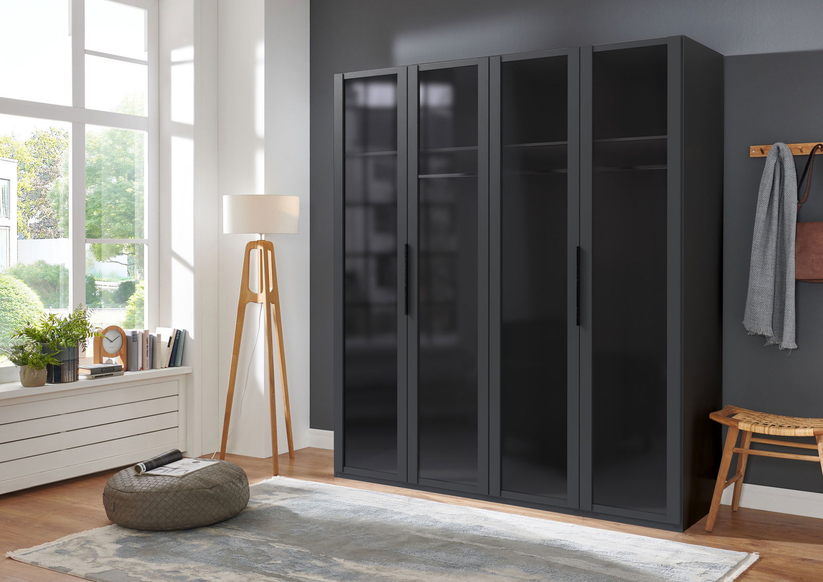 Moderner schwarzer Drehtürenschrank mit vier Türen, frontal fotografiert in einem hellen Raum mit großen Fenstern.