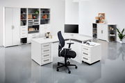 Moderne Büroumgebung mit weißem Eckschreibtisch, Rollcontainer und Bürostuhl, frontal betrachtet.