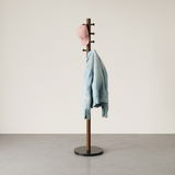 Moderner Garderobenständer aus Holz mit schwarzem Sockel, von der Seite gesehen, mit einer blauen Jeansjacke und einer rosa Kappe behängt.