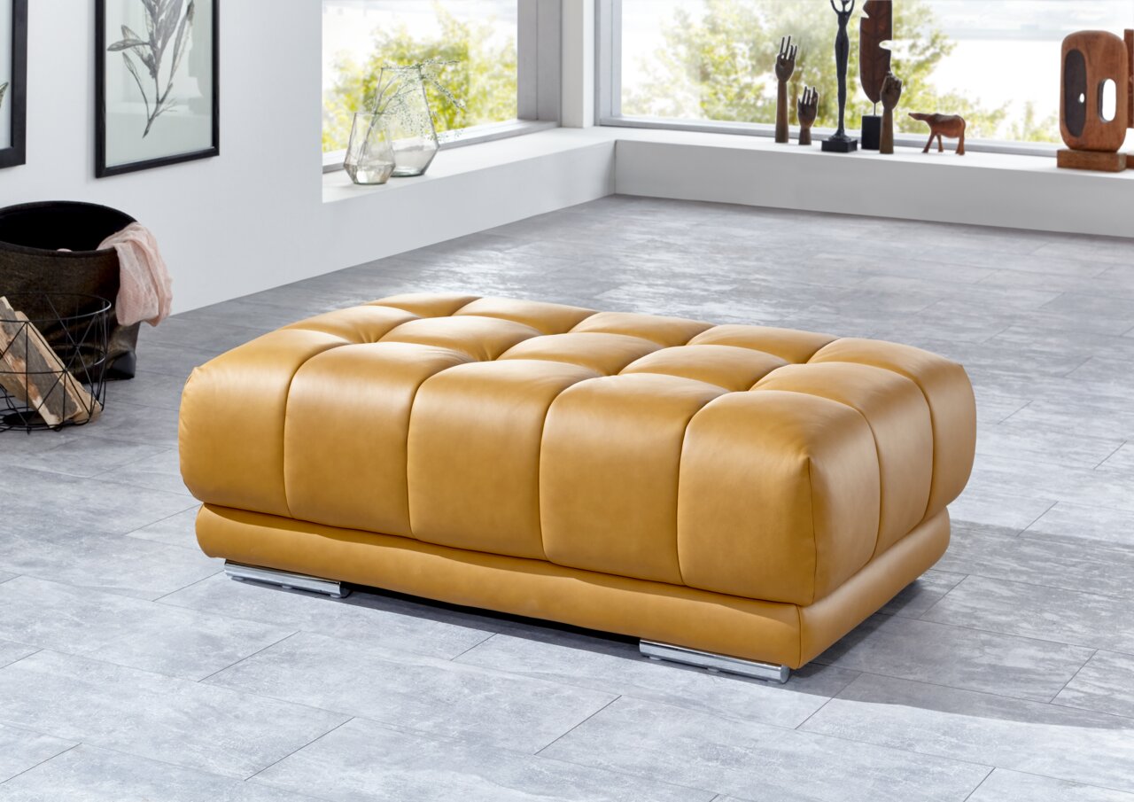 XXL-Hocker in hellbraunem Leder, seitliche Perspektive, in modernem Wohnzimmer mit großen Fenstern.