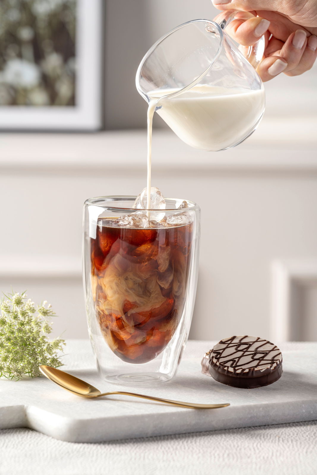 Doppelwandbecher mit Eiskaffee und Milch, von der Seite fotografiert, auf einem Tisch mit einem Keks und einem goldenen Löffel.