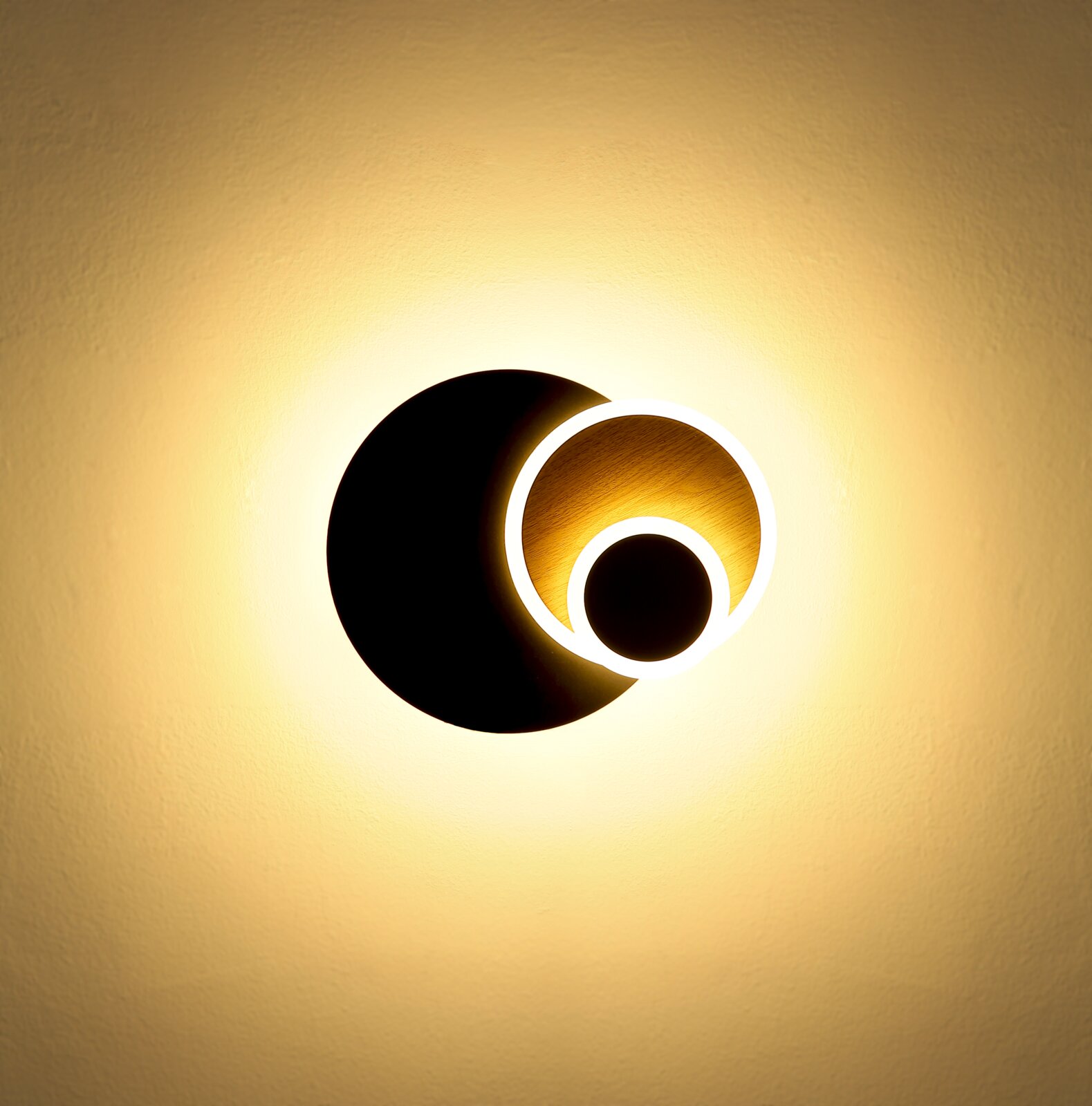 Moderne runde LED-Wandleuchte in Schwarz mit goldfarbener Innenfläche und leuchtenden Ringelementen, warmweißes indirektes Licht an heller Wand, Frontalansicht von vorn