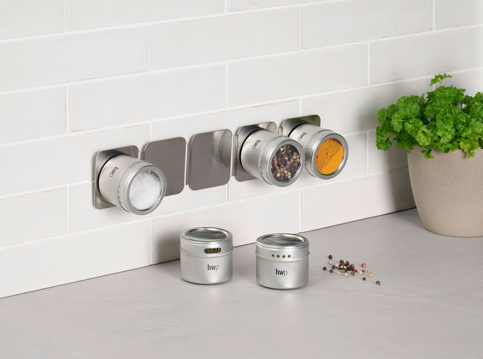 Homeware Gewürzdosenset 6-tlg. Gewürzdosenset an der Wand montiert, mit drei Dosen gefüllt mit Salz, Pfeffer und Kurkuma, und zwei Dosen auf der Arbeitsfläche. Perspektive von vorne.