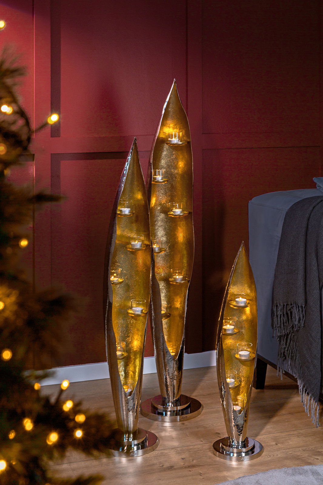Drei elegante, goldene Kerzenhalter in Blattform mit Teelichtern, stehend auf einem Holzboden vor einer roten Wand. Perspektive von vorne.