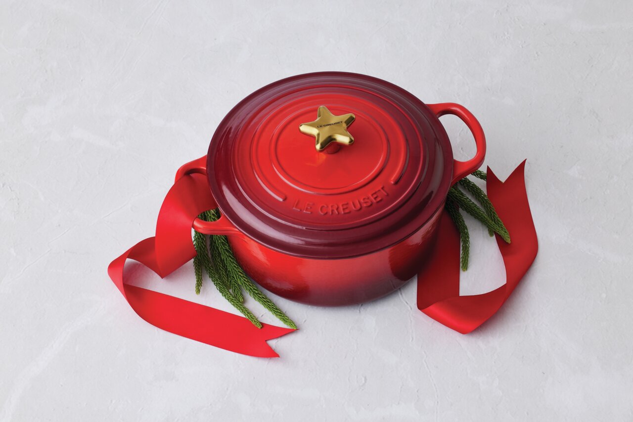 LE CREUSET Deckelknauf Stern Rote Le Creuset Kasserolle mit goldenem Stern-Deckelknopf, dekoriert mit rotem Band und Tannenzweigen, von oben fotografiert.