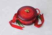 Rote Le Creuset Kasserolle mit goldenem Stern-Deckelknopf, dekoriert mit rotem Band und Tannenzweigen, von oben fotografiert.