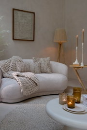 Gemütliche Wohnzimmer-Szene mit einer beigen Couch, dekoriert mit Kissen und einer Strickdecke, im Vordergrund ein runder Couchtisch mit Kerzen, seitliche Perspektive.