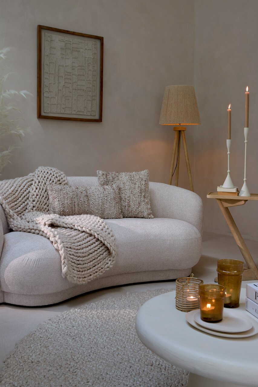Gemütliche Wohnzimmer-Szene mit einer beigen Couch, dekoriert mit Kissen und einer Strickdecke, im Vordergrund ein runder Couchtisch mit Kerzen, seitliche Perspektive.