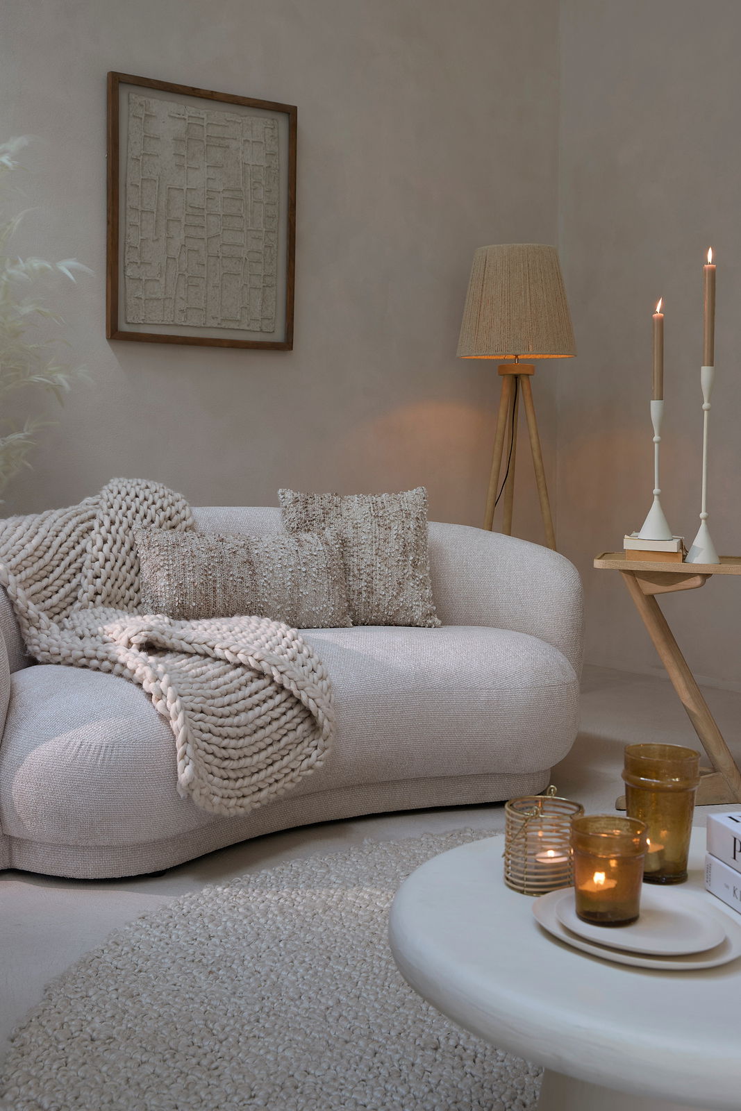 Gemütliche Wohnzimmer-Szene mit einer beigen Couch, dekoriert mit Kissen und einer Strickdecke, im Vordergrund ein runder Couchtisch mit Kerzen, seitliche Perspektive.