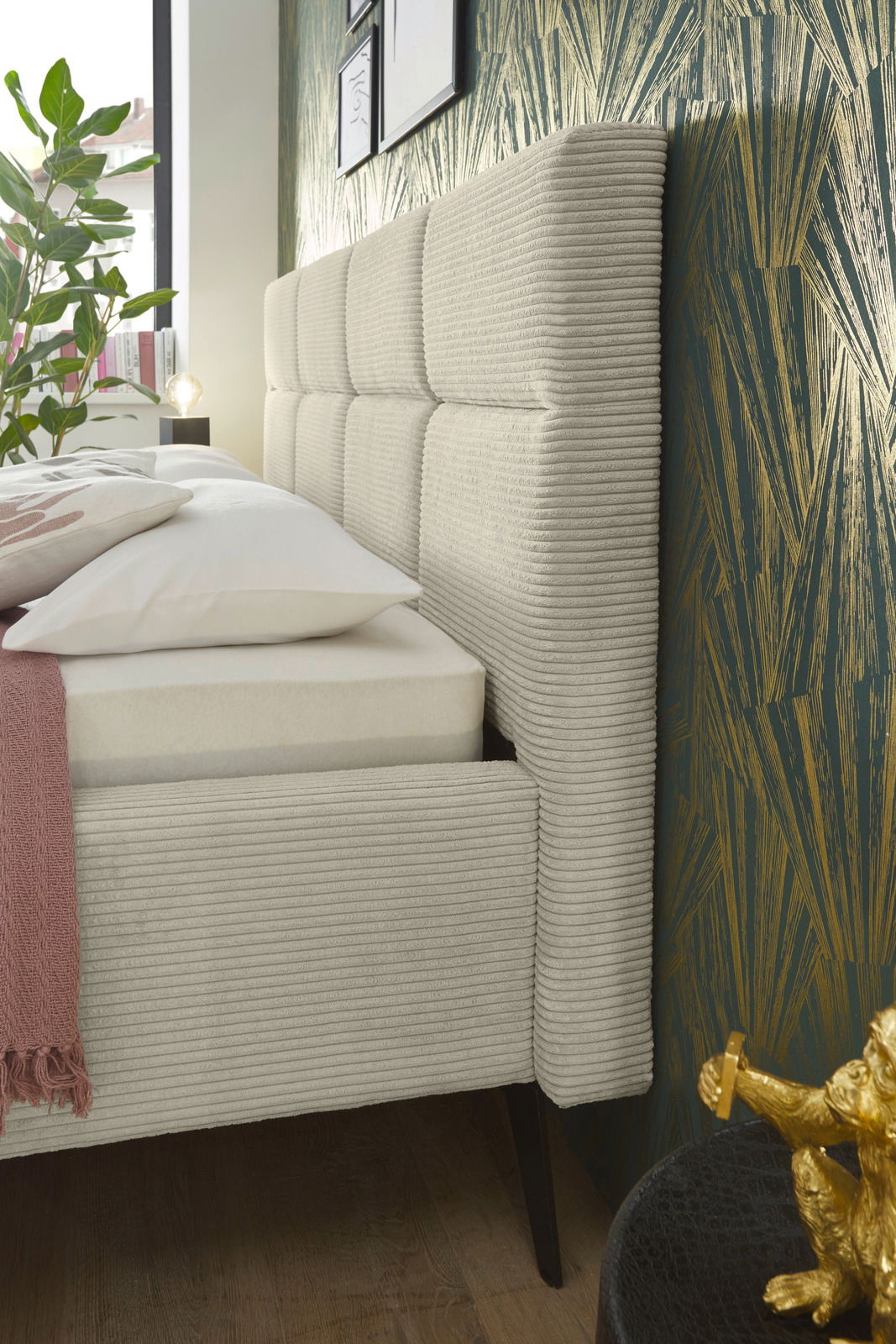 Seitliche Nahaufnahme eines modernen Polsterbetts mit gestepptem Kopfteil und eleganten Stoffbezügen, dekoriert mit Kissen und Decke, vor einer stilvollen Tapete.