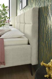 Seitliche Nahaufnahme eines modernen Polsterbetts mit gestepptem Kopfteil und eleganten Stoffbezügen, dekoriert mit Kissen und Decke, vor einer stilvollen Tapete.