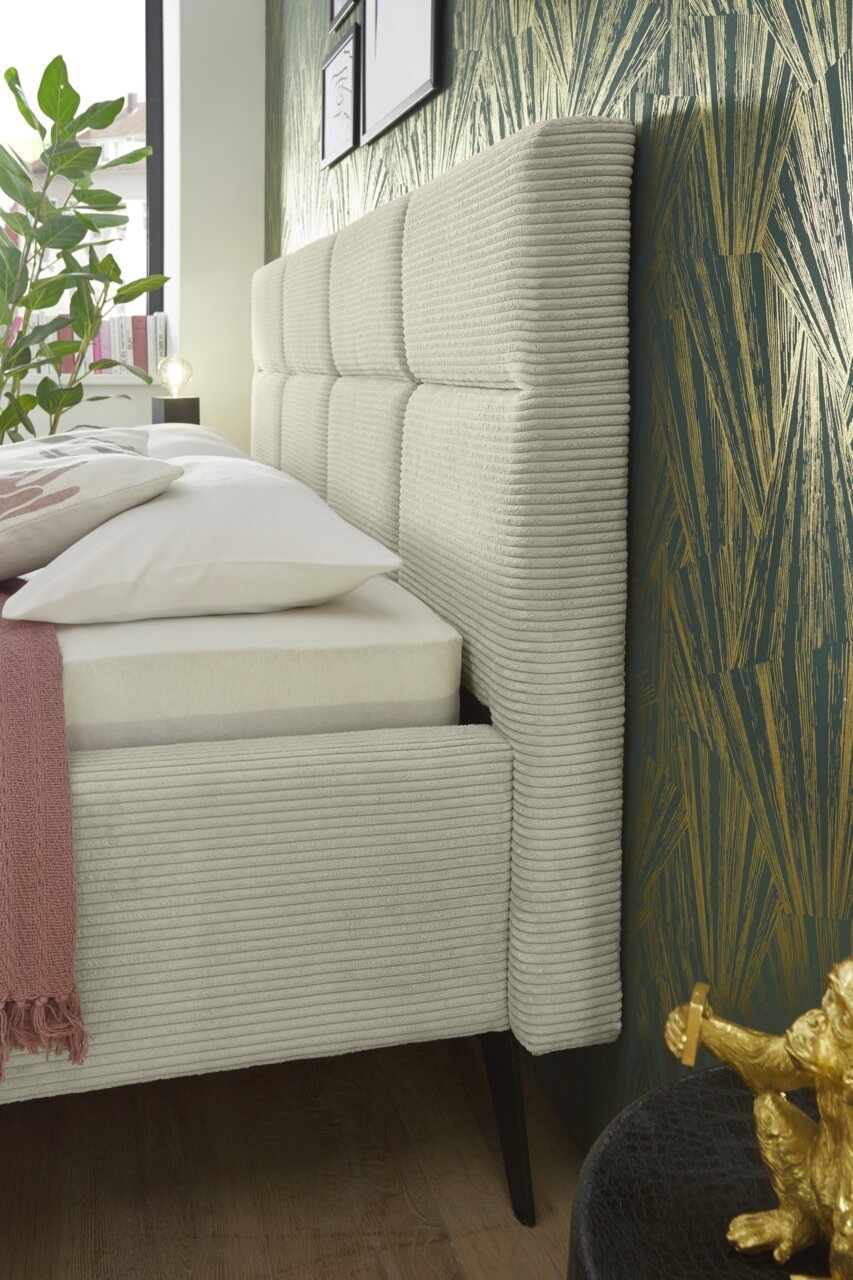 Seitliche Nahaufnahme eines modernen Polsterbetts mit gestepptem Kopfteil und eleganten Stoffbezügen, dekoriert mit Kissen und Decke, vor einer stilvollen Tapete.