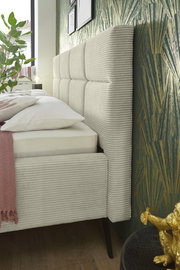 Seitliche Nahaufnahme eines modernen Polsterbetts mit gestepptem Kopfteil und eleganten Stoffbezügen, dekoriert mit Kissen und Decke, vor einer stilvollen Tapete.