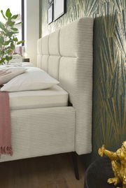 Seitliche Nahaufnahme eines modernen Polsterbetts mit gestepptem Kopfteil und eleganten Stoffbezügen, dekoriert mit Kissen und Decke, vor einer stilvollen Tapete.