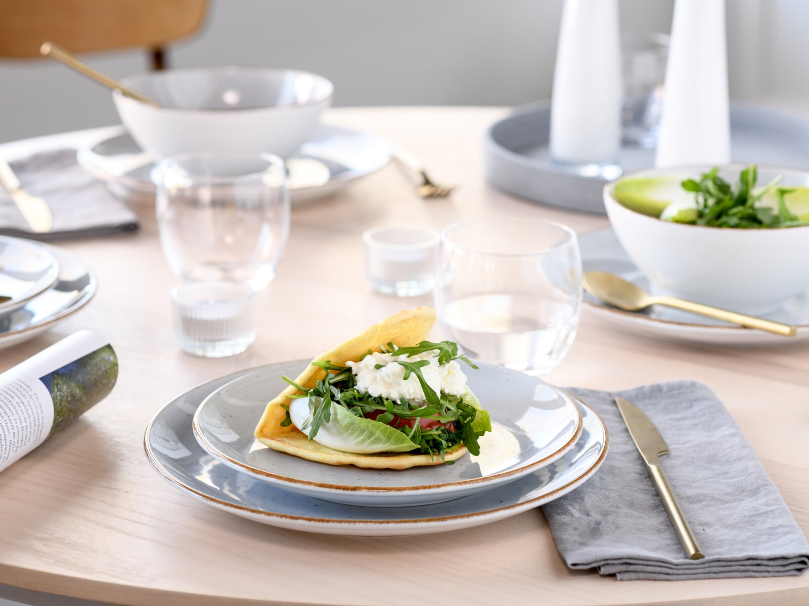 Schlicht gedeckter Esstisch mit grauen Keramiktellern, Wassergläsern und einem gefüllten Fladenbrot mit Rucola und Käse auf einem Teller, aufgenommen aus leicht erhöhter schräger Vorderansicht.