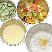 Vier Küchenschüsseln aus Edelstahl, gefüllt mit Gurkenscheiben, Obstsalat, Teigflüssigkeit und Mehl mit Butter, aus der Vogelperspektive.