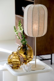 Beige tafellamp met ovale, geribde stoffen kap en slanke voet, vanuit schuin vooraanzicht gefotografeerd naast amberkleurige glazen vazen.