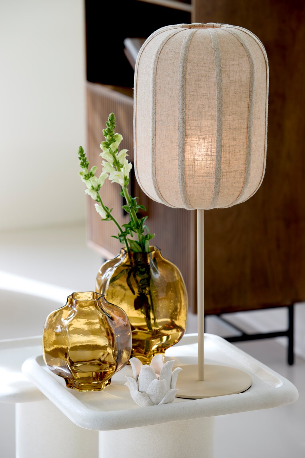 Beige tafellamp met ovale, geribde stoffen kap en slanke voet, vanuit schuin vooraanzicht gefotografeerd naast amberkleurige glazen vazen.