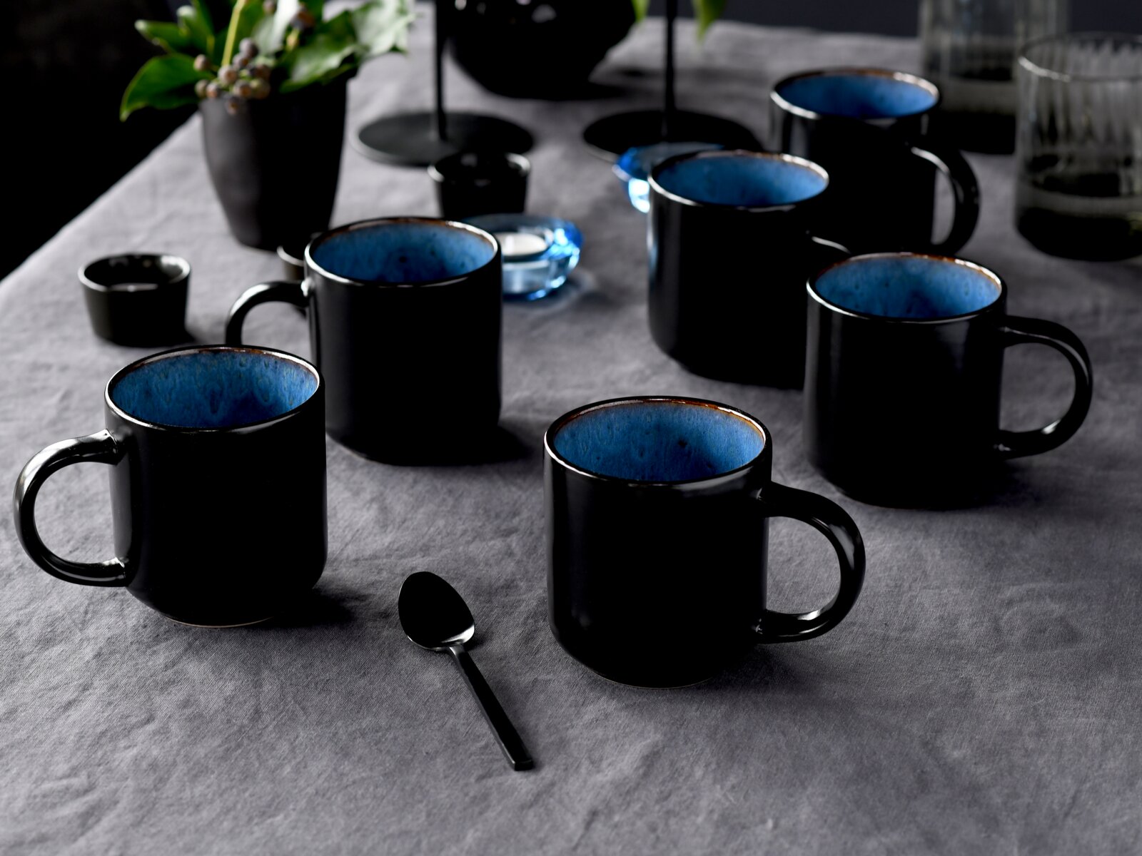 Schwarze Keramik-Kaffeebecher mit blau glasierter Innenseite auf grauem Tischtuch, schräg von vorne in leichter Aufsicht fotografiert
