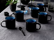Schwarze Keramik-Kaffeebecher mit blau glasierter Innenseite auf grauem Tischtuch, schräg von vorne in leichter Aufsicht fotografiert
