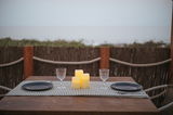 Drei leuchtende Outdoor-Kerzen auf einem gedeckten Tisch mit zwei Gläsern und Tellern, im Freien mit Blick auf das Meer, aus der Perspektive eines sitzenden Betrachters.