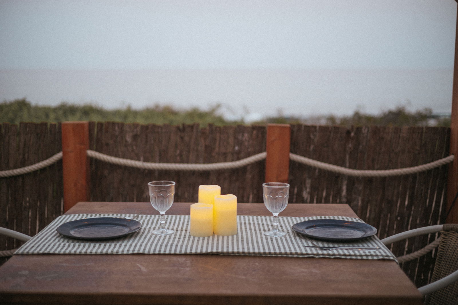 Drei leuchtende Outdoor-Kerzen auf einem gedeckten Tisch mit zwei Gläsern und Tellern, im Freien mit Blick auf das Meer, aus der Perspektive eines sitzenden Betrachters.