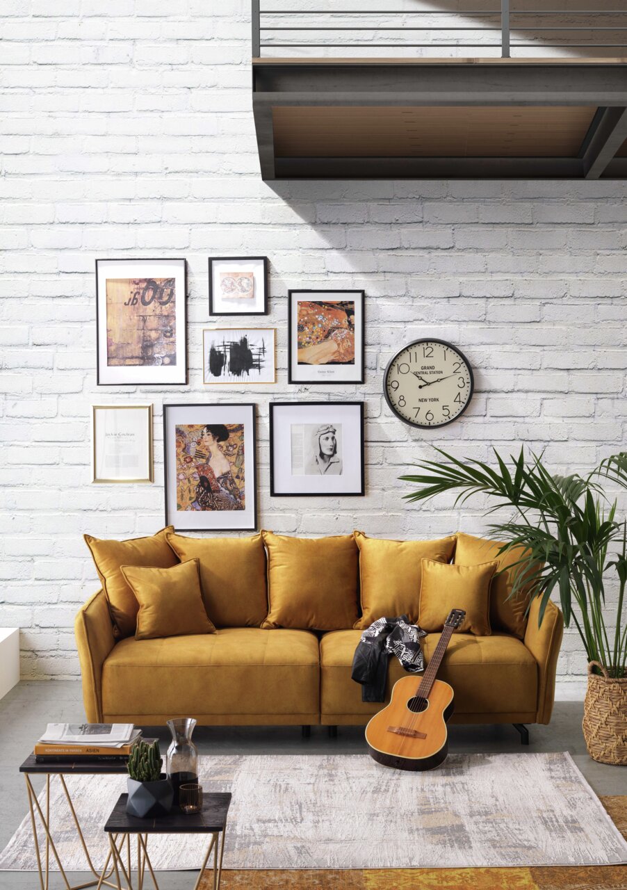Gemütliches Big-Sofa in Senfgelb vor einer weißen Backsteinwand, dekoriert mit Kunstwerken und einer Uhr. Perspektive: Frontalansicht.