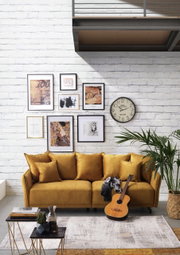 Gemütliches Big-Sofa in Senfgelb vor einer weißen Backsteinwand, dekoriert mit Kunstwerken und einer Uhr. Perspektive: Frontalansicht.