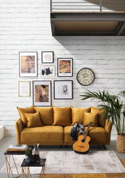 Gemütliches Big-Sofa in Senfgelb vor einer weißen Backsteinwand, dekoriert mit Kunstwerken und einer Uhr. Perspektive: Frontalansicht.