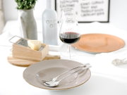 Heller, modern gedeckter Esstisch in leichter Schrägansicht von vorn: beiger Keramikteller mit Löffel und Gabel, daneben Rotweinglas, Käsehobel mit Käse, orangefarbener Teller sowie Vase und Flasche im Hintergrund.