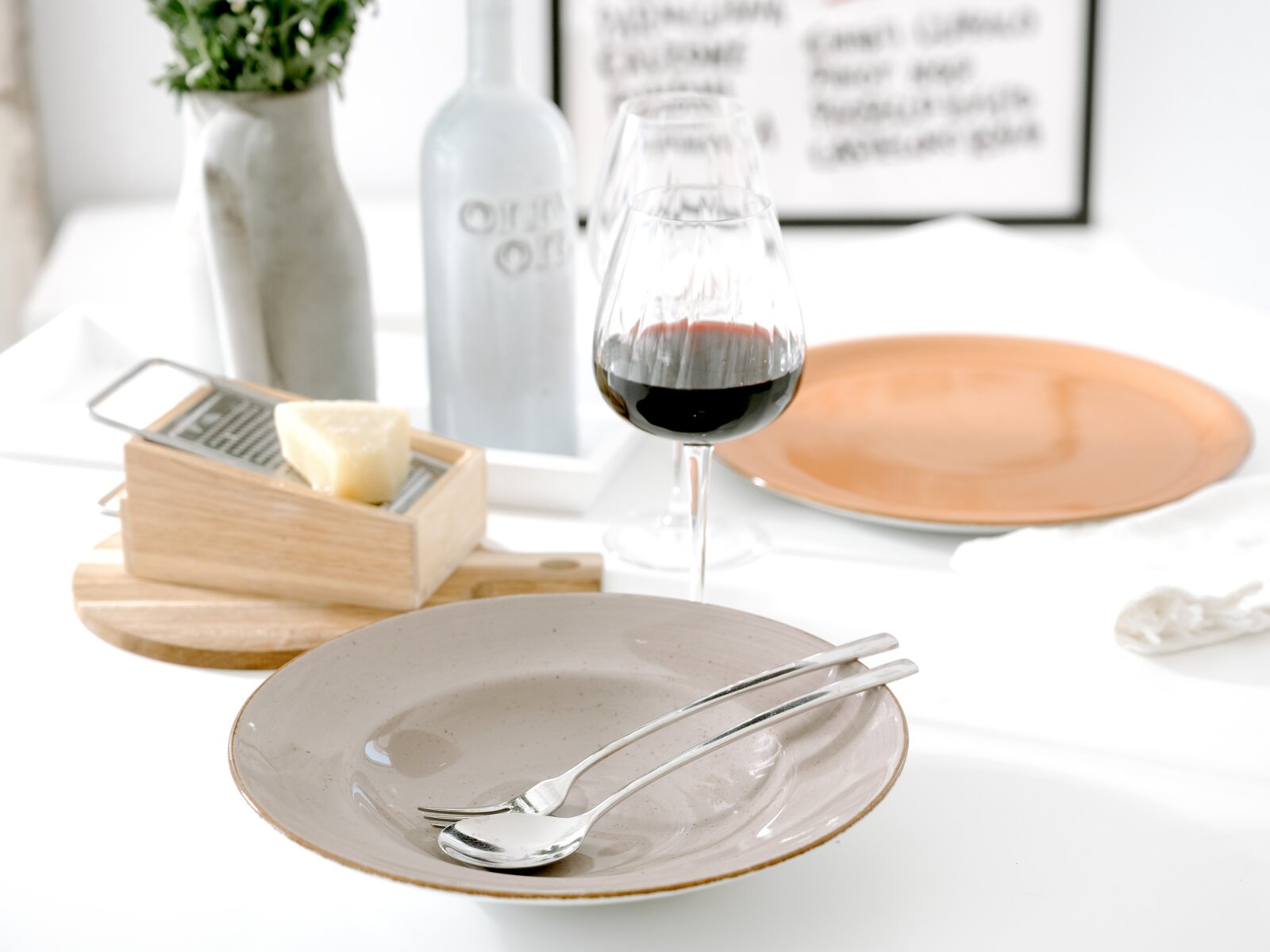Heller, modern gedeckter Esstisch in leichter Schrägansicht von vorn: beiger Keramikteller mit Löffel und Gabel, daneben Rotweinglas, Käsehobel mit Käse, orangefarbener Teller sowie Vase und Flasche im Hintergrund.