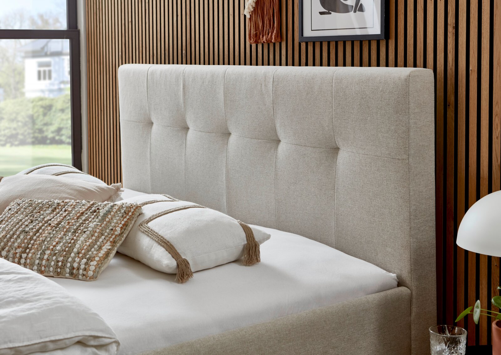 Beige boxspring met doorgestikt hoofdeinde, gezien vanaf de zijkant, versierd met kussens in een moderne slaapkamer.