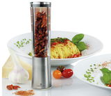 Silberne Gewürzmühle mit transparentem Behälter, gefüllt mit getrockneten Chilis, im Vordergrund, umgeben von einem Teller mit Spaghetti, Knoblauch, Kirschtomaten und Parmesan. Perspektive: Frontalansicht.