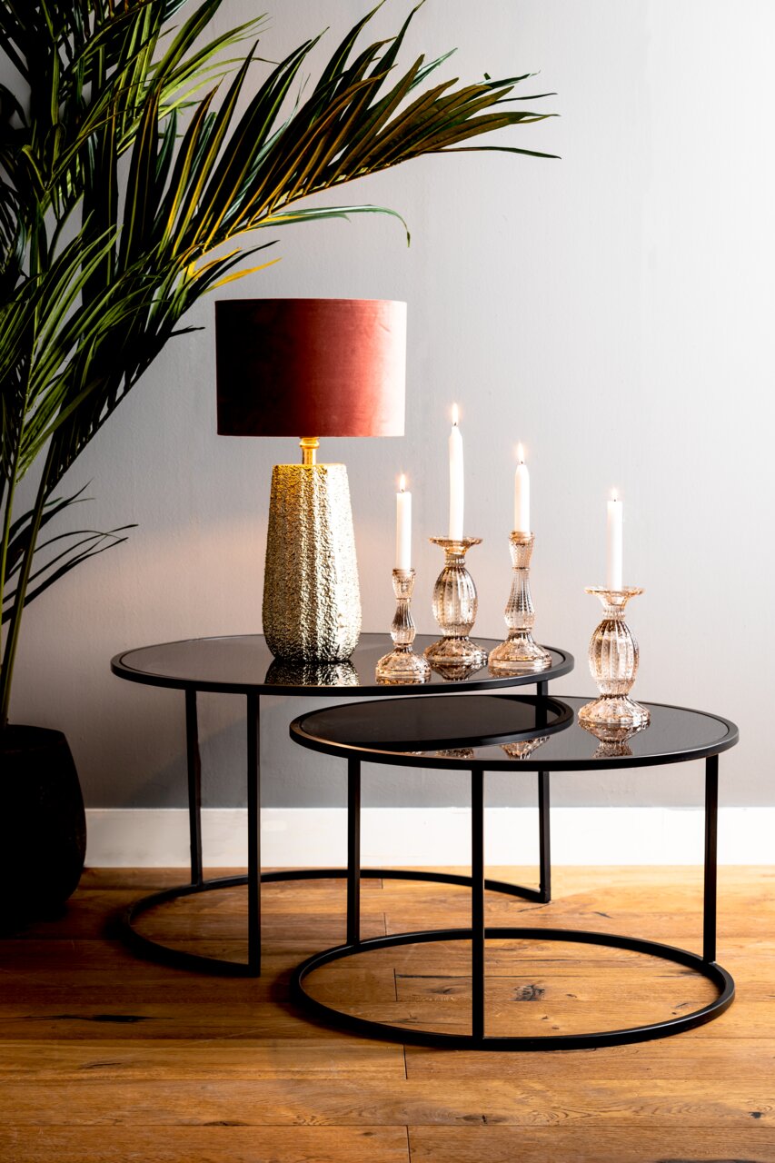 Zwei runde Couchtische in verschiedenen Größen mit schwarzer Glasplatte und Metallrahmen, dekoriert mit einer Tischlampe und Kerzenhaltern, aus seitlicher Perspektive fotografiert.