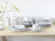 Weißes Porzellan-Geschirrset mit stapelbaren rechteckigen Tellern/Platten und Kaffeetassen auf hellem Holztisch vor weißer Wand, perspektivisch in leichter Schrägansicht von vorne.