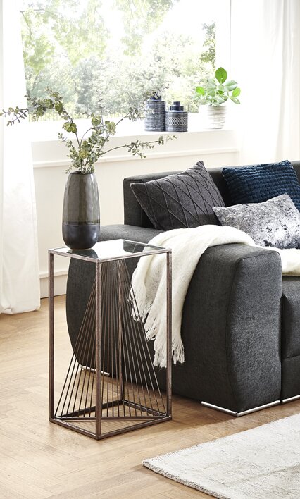 Moderner Beistelltisch mit Metallrahmen und schwarzer Faden-/Schnur-Optik neben grauem Sofa, darauf eine dunkle Vase; Perspektive: leicht schräg von vorne auf Augenhöhe.