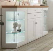 Weiße Anrichte/Sideboard im Landhausstil mit Holzplatte, Glasvitrine links und Schubladen rechts, seitliche Perspektive von vorne links in hellem Wohnraum