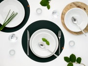 Modernes Tischgedeck mit weißen Tellern und Schüsseln auf dunklen runden Platzsets, Besteck, Wassergläsern und Teelichtern, dekoriert mit Kräutern und grünen Blättern, Draufsicht (Top-Down) auf weißem Hintergrund