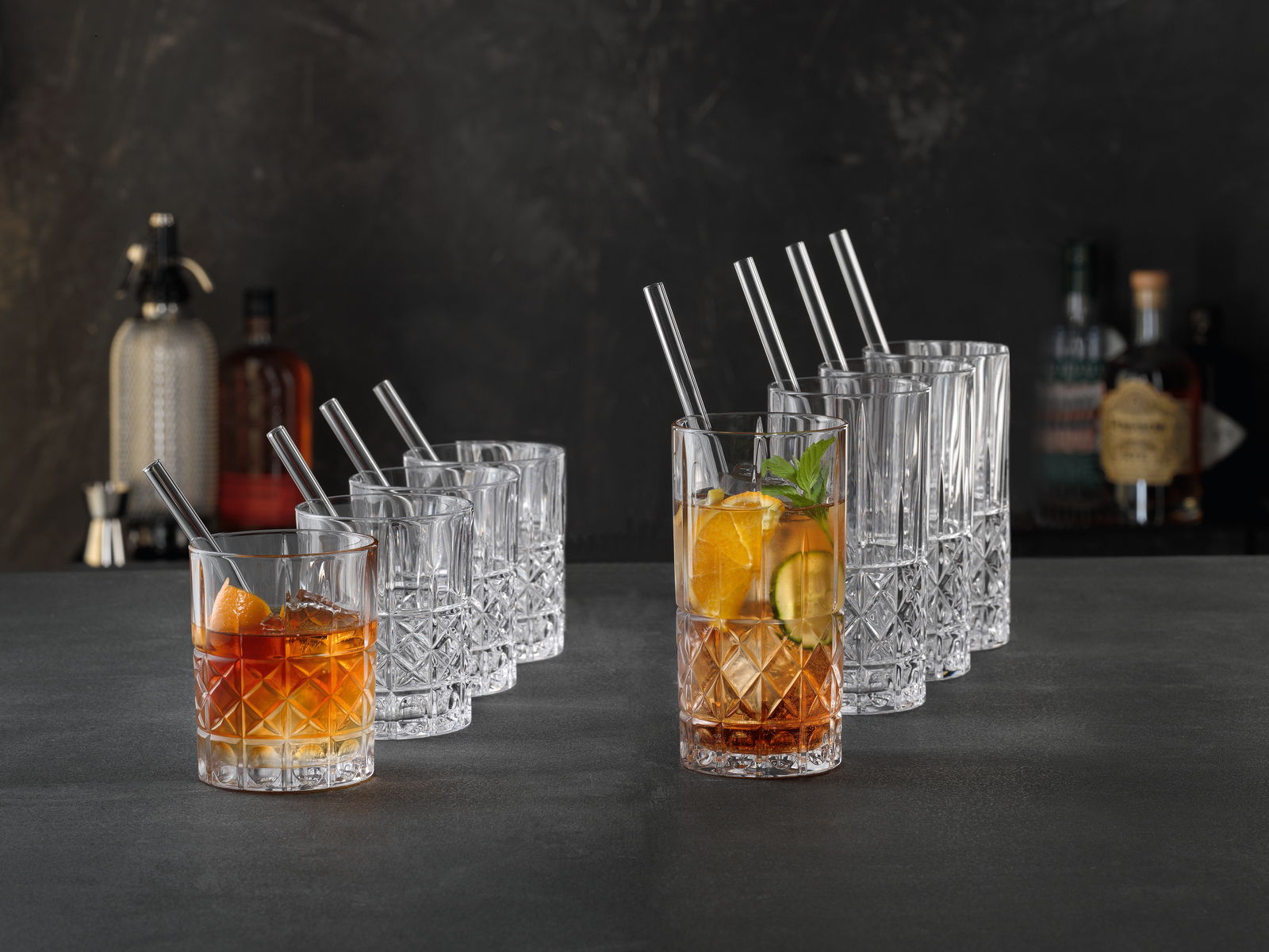 Ein Set von eleganten Kristallgläsern, bestehend aus acht niedrigen und acht hohen Gläsern, auf einer dunklen Oberfläche präsentiert. Die Gläser sind teilweise mit Getränken und Eiswürfeln gefüllt, dekoriert mit Orangenscheiben und Minzblättern. Perspekti