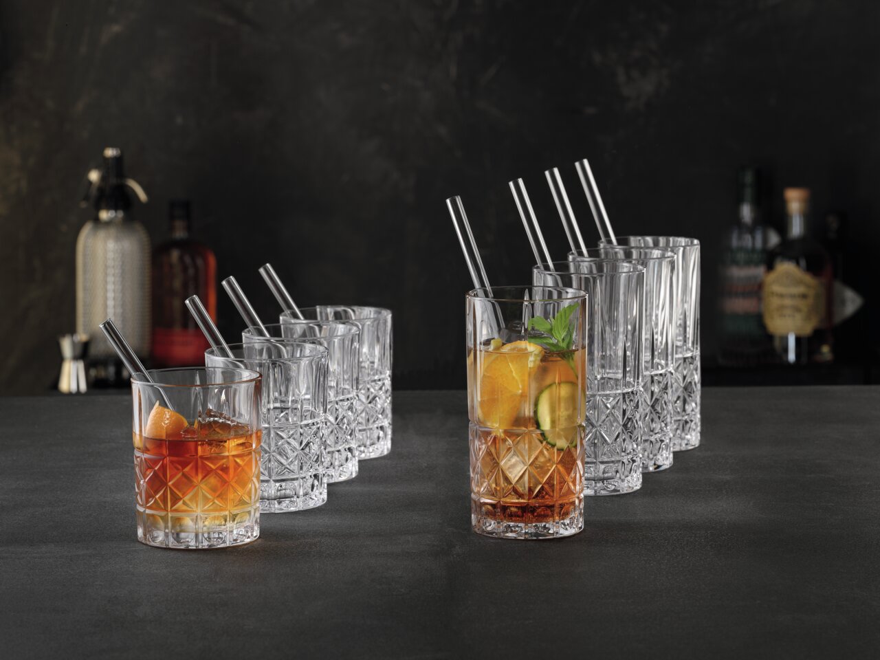 Ein Set von eleganten Kristallgläsern, bestehend aus acht niedrigen und acht hohen Gläsern, auf einer dunklen Oberfläche präsentiert. Die Gläser sind teilweise mit Getränken und Eiswürfeln gefüllt, dekoriert mit Orangenscheiben und Minzblättern. Perspekti