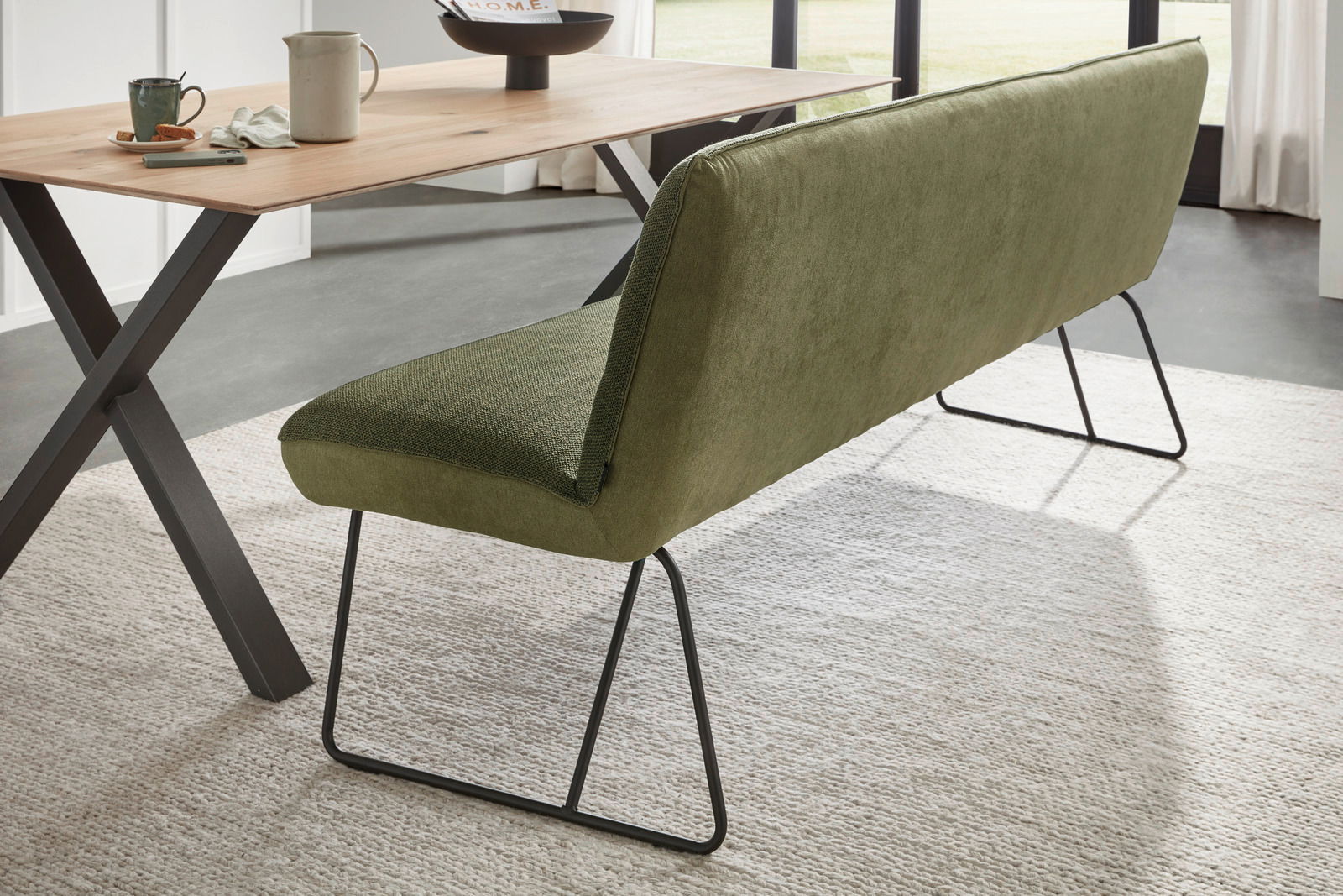 Grüne Polsterbank mit Metallbeinen, seitliche Perspektive neben einem Holztisch.