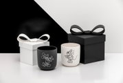 Zwei matte Kerzenhalter/Becher in Schwarz und Weiß mit Linienillustrationen einer Mausfigur, vorne in leicht seitlicher Perspektive fotografiert, daneben Geschenkboxen mit Schleifen in Schwarz und Weiß vor zweifarbigem Hintergrund.