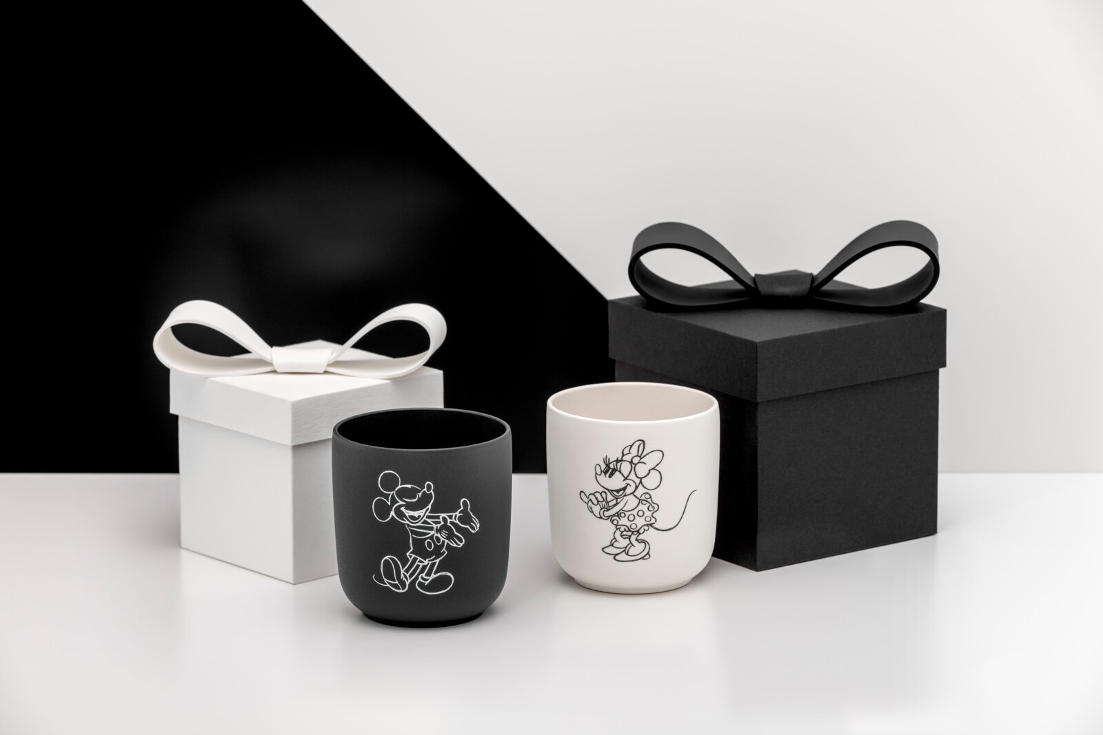 Zwei matte Kerzenhalter/Becher in Schwarz und Weiß mit Linienillustrationen einer Mausfigur, vorne in leicht seitlicher Perspektive fotografiert, daneben Geschenkboxen mit Schleifen in Schwarz und Weiß vor zweifarbigem Hintergrund.