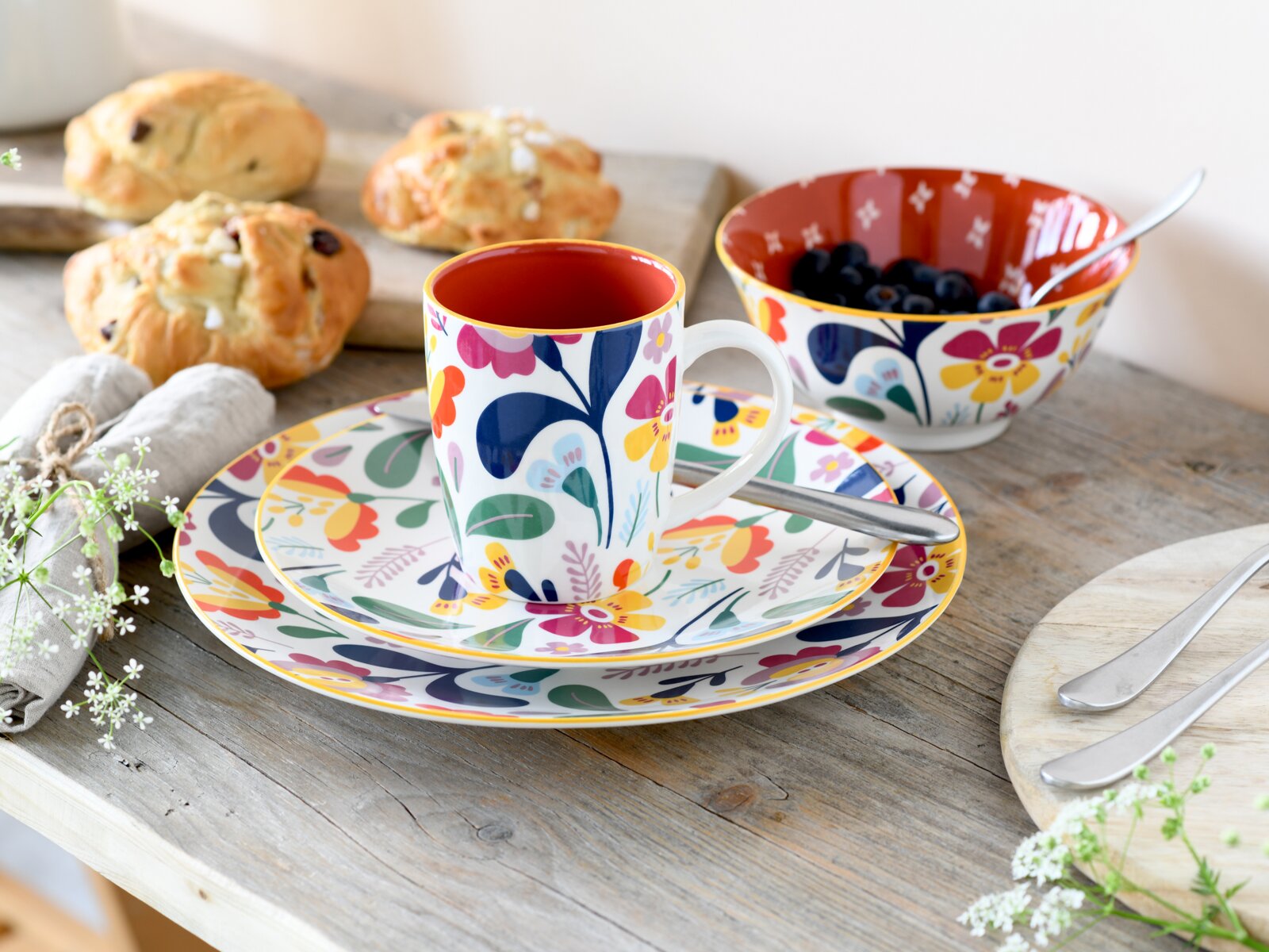 Buntes, florales Geschirr-Set mit Tasse auf Untertasse und großem Teller sowie Schale mit Blaubeeren und Löffel auf rustikalem Holztisch, fotografiert aus leicht erhöhter schräger Perspektive (Dreiviertelansicht).