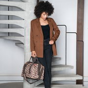 Frau in braunem Blazer und schwarzer Hose steht auf einer Treppe und hält eine Tasche mit Leopardenmuster in der Hand, seitliche Perspektive.